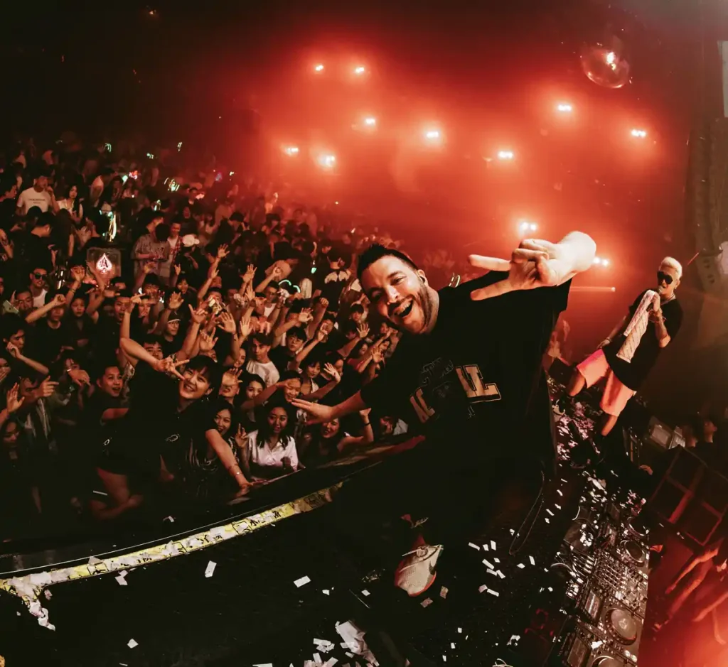Amid the electrifying atmosphere of one of the Top 10 Best Nightclubs in China, a DJ performs on stage with a cheering crowd illuminated by red lights. The DJ is making a peace sign while standing behind the turntables.
