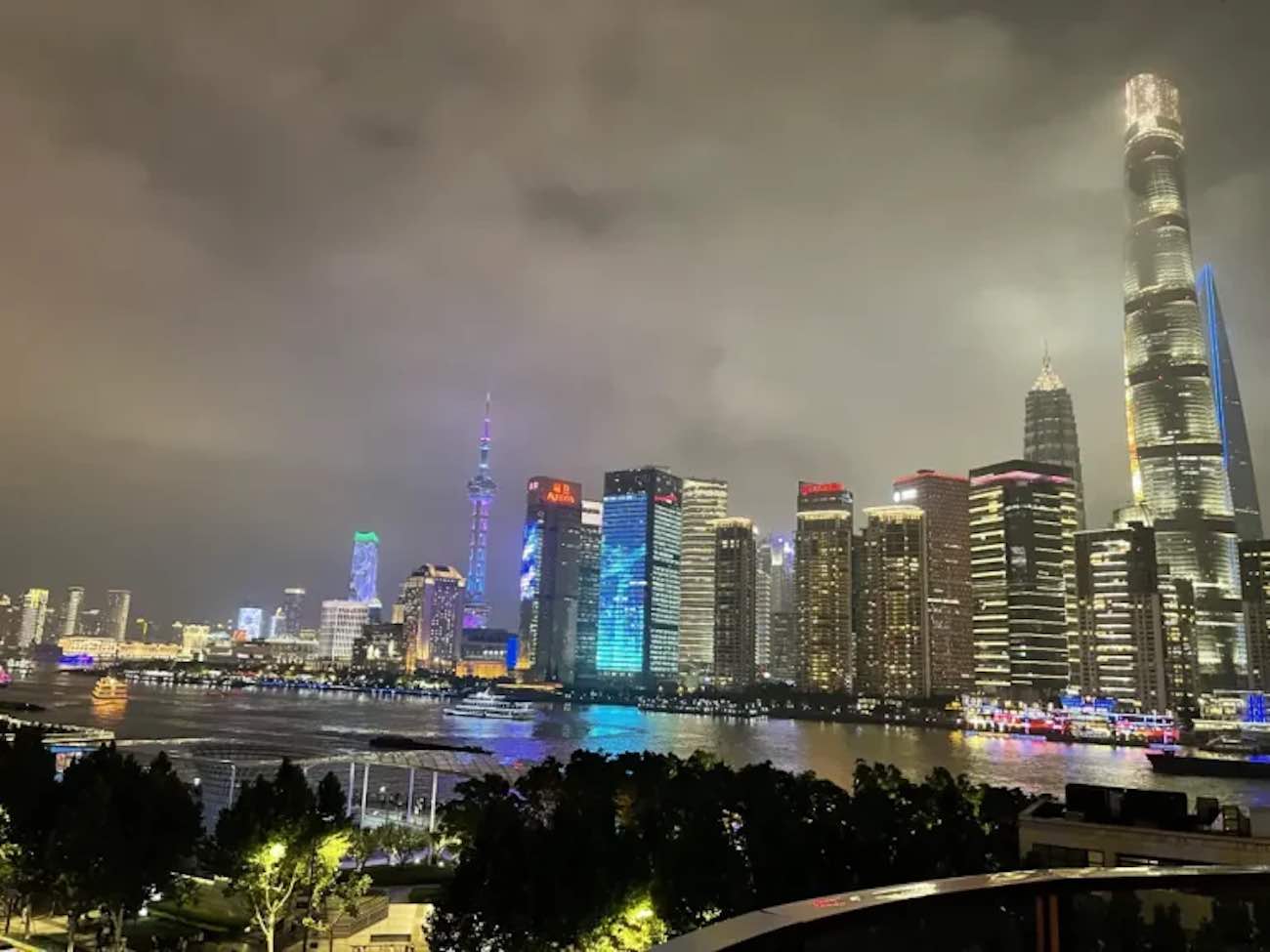 Shanghai 's nachts: 24+ beste dingen om te doen & 8 tips van experts 15 Shanghai 's nachts Een nachtelijk uitzicht op de skyline van een stad met verlichte wolkenkrabbers langs de waterkant, waarbij het hoogste gebouw verlicht en prominent aanwezig is in het rechtergedeelte van de afbeelding onder een bewolkte hemel, wat doet denken aan een scène uit een van de 10 beste nachtclubs in China.