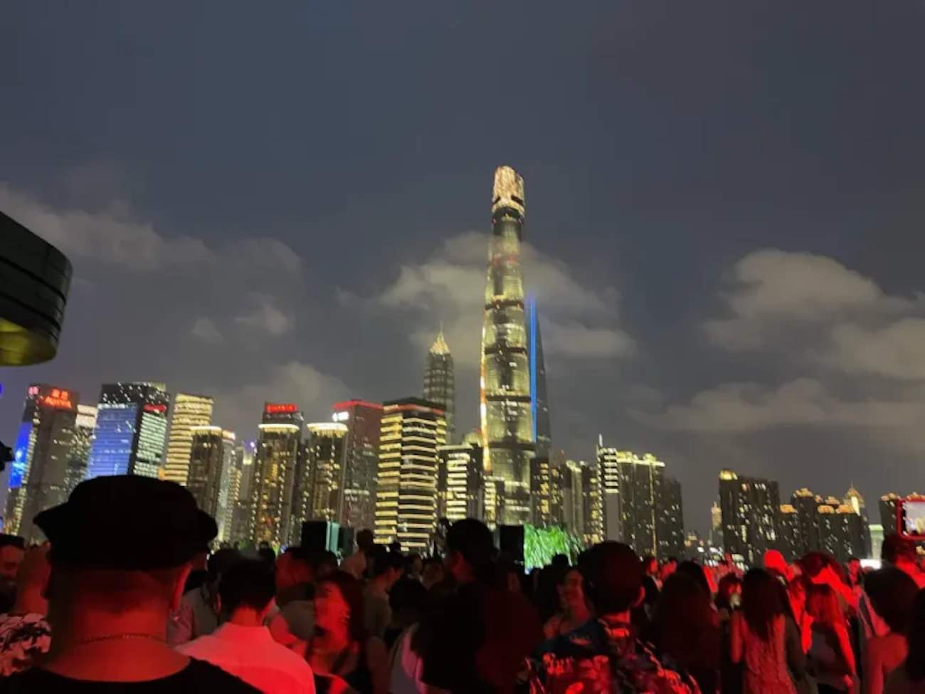 Shanghai at night: best nightlife handbook (2025) 53 renting a car in okinawa A crowd gathers at night with illuminated skyscrapers, including a tall, brightly lit tower, in the background under a cloudy sky. Among the vibrant scene are some of the top 10 best nightclubs in china, adding to the lively atmosphere.