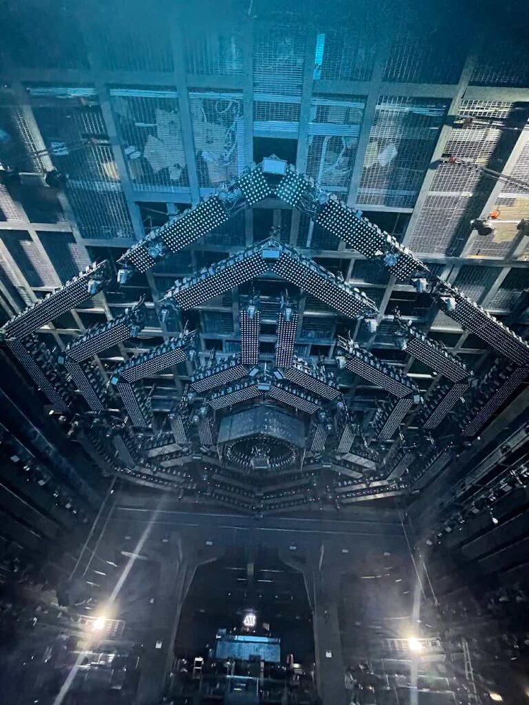 A view of the interior of a large, modern concert venue with a geometric, illuminated ceiling design and light fixtures.