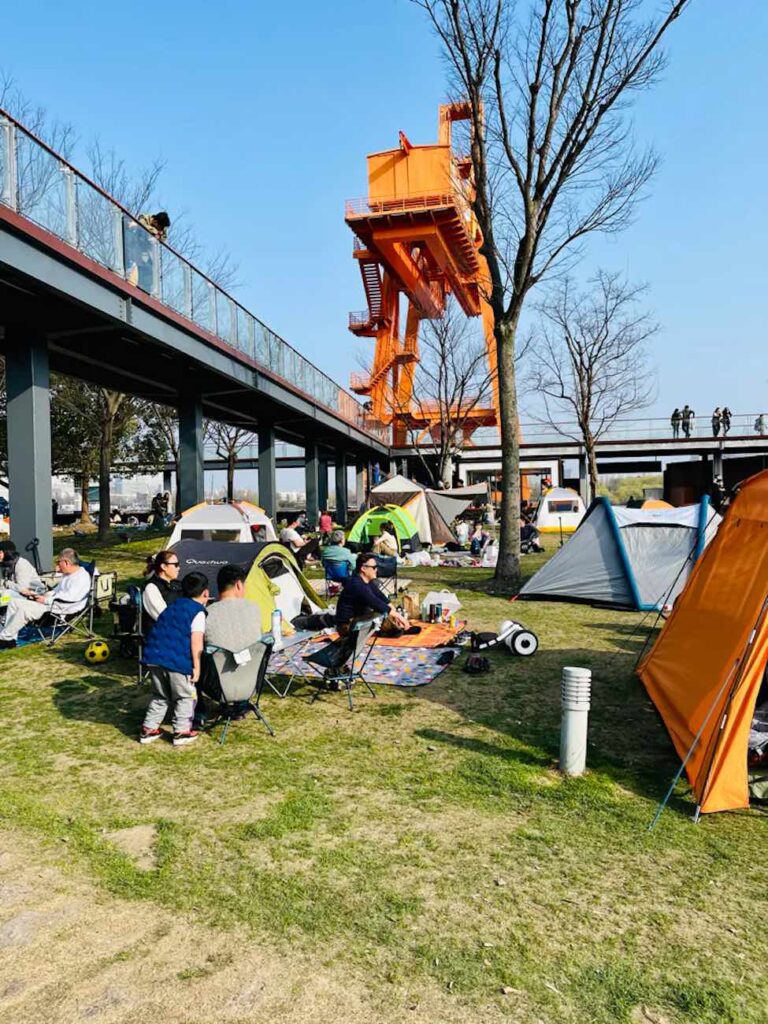 57 things to do in shanghai: the ultimate handbook 22 things to do in shanghai People camping in a grassy area under a bridge at xu hui river side in shanghai with several tents and an orange industrial structure in the background, showing unique outdoor things to do in shanghai where campers on folding chairs make for unbelievable photos. -copyright-moving-jack. Com