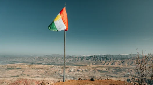 Is iraqi kurdistan and erbil safe? Complete safety guide (2025) 11 A flag on a pole stands proudly on a mountain in kurdistan, overlooking a wide valley with distant hills under a clear blue sky.