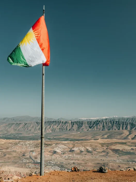 Is iraqi kurdistan and erbil safe? Complete safety guide (2025) 12 A flag on a pole stands proudly on a mountain in kurdistan, overlooking a wide valley with distant hills under a clear blue sky.