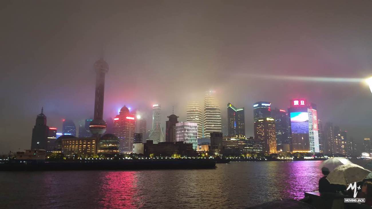 Een nachtelijk stadsgezicht van de skyline van Shanghai, met hoge gebouwen en kleurrijke lichtjes die door de dikke mist boven de rivier schijnen en in het water reflecteren – een adembenemend tafereel dat zowel de aantrekkingskracht als de complexiteit van het leven in het centrum van Shanghai vastlegt. -Copyright-moving-jack.com