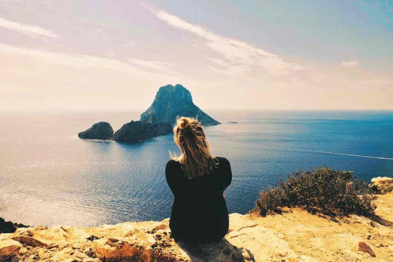 A person with blonde hair sits on a rocky cliff, facing a distant island in the calm ocean under a clear sky, taking a moment to relax on ibiza.