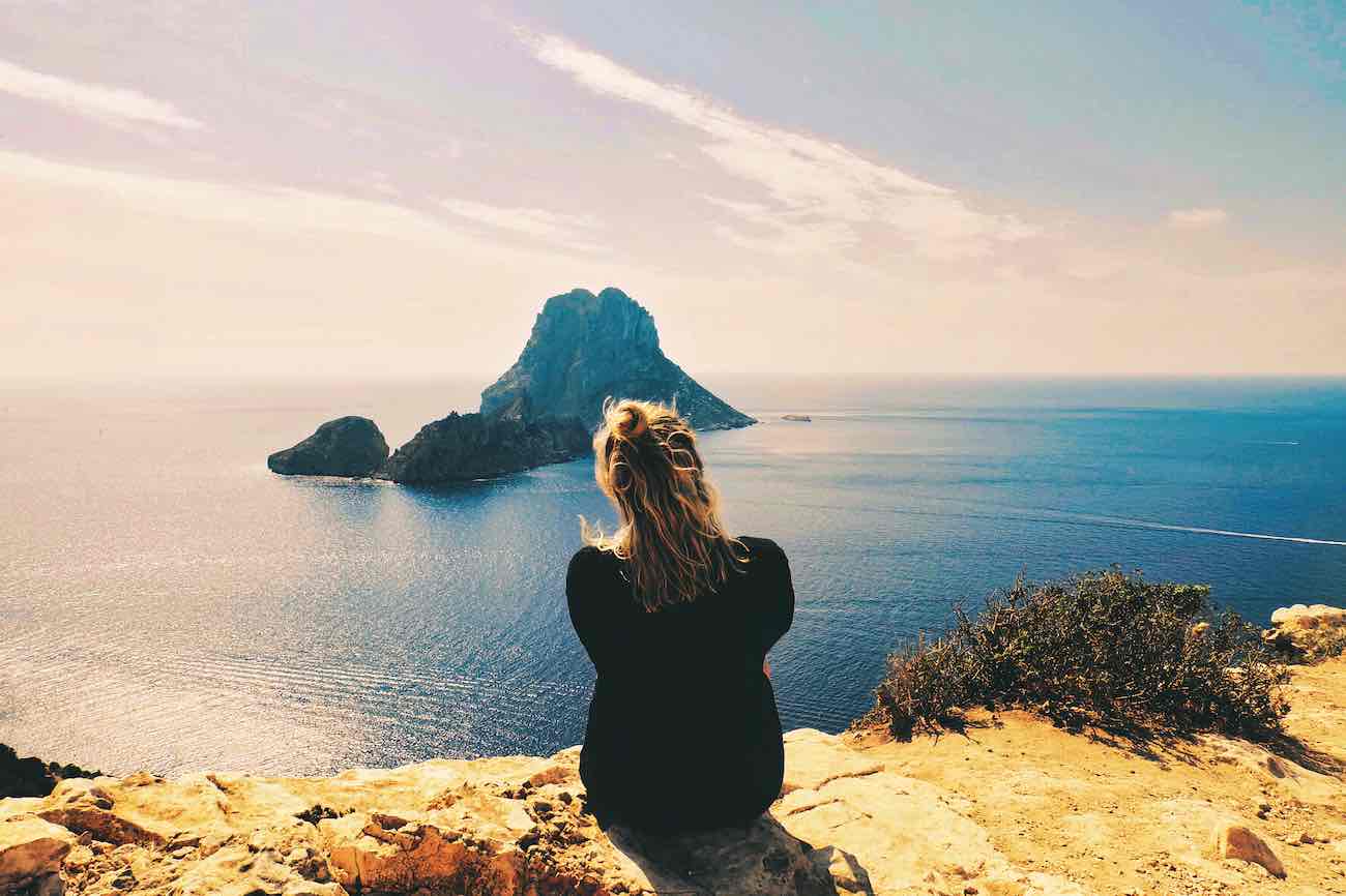 Een persoon met blond haar zit op een rotsachtige klif, met uitzicht op een verafgelegen eiland in de kalme oceaan onder een heldere hemel, en neemt een moment om te ontspannen op Ibiza.