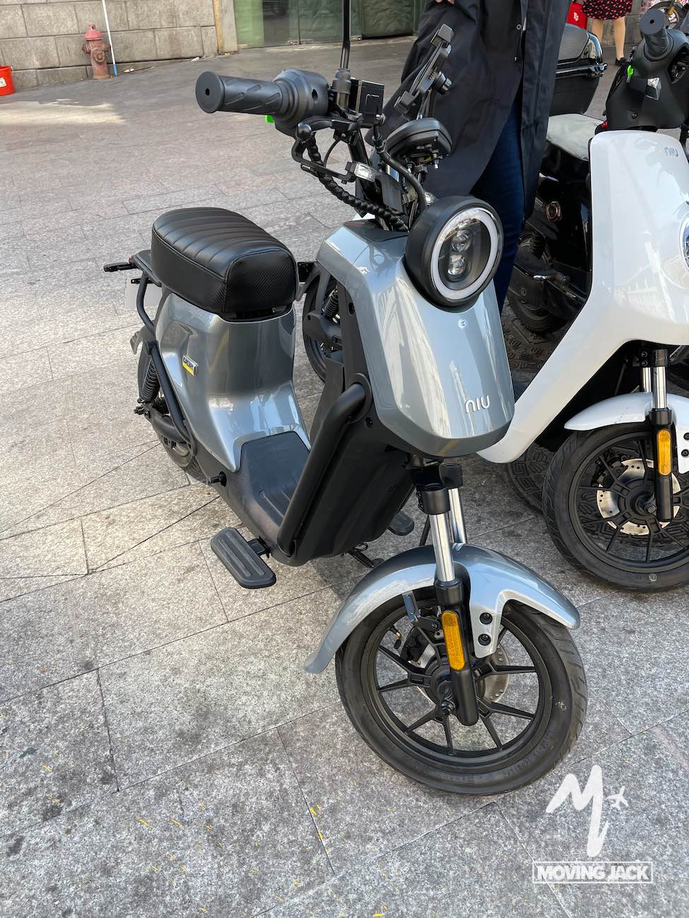 Een moderne grijze Niu elektrische scooter, geparkeerd op een stoep in de stad, geeft de sfeer weer van het leven in het centrum van Shanghai, met het "MOVING JACK"-logo op de grond en een persoon die erachter staat. -Copyright-moving-jack.com