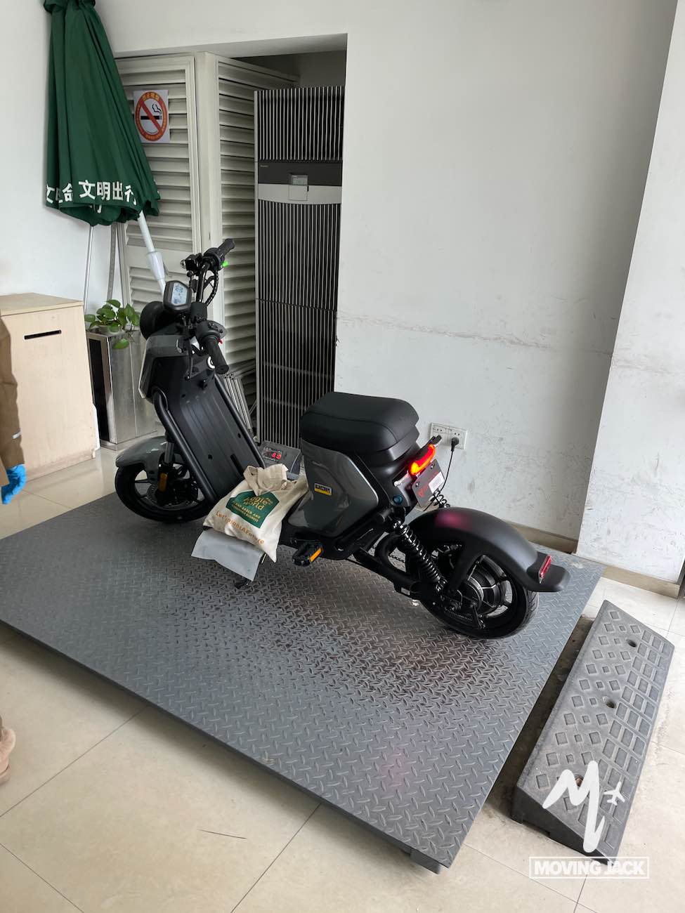 Een zwarte elektrische scooter staat geparkeerd op een verhoogd metalen platform binnen, wat de gemakken van het wonen in het centrum van Shanghai weerspiegelt. Aan het stuur hangt een tas, met op de achtergrond een groene paraplu en een betegelde vloer en witte muren. -Copyright-moving-jack.com