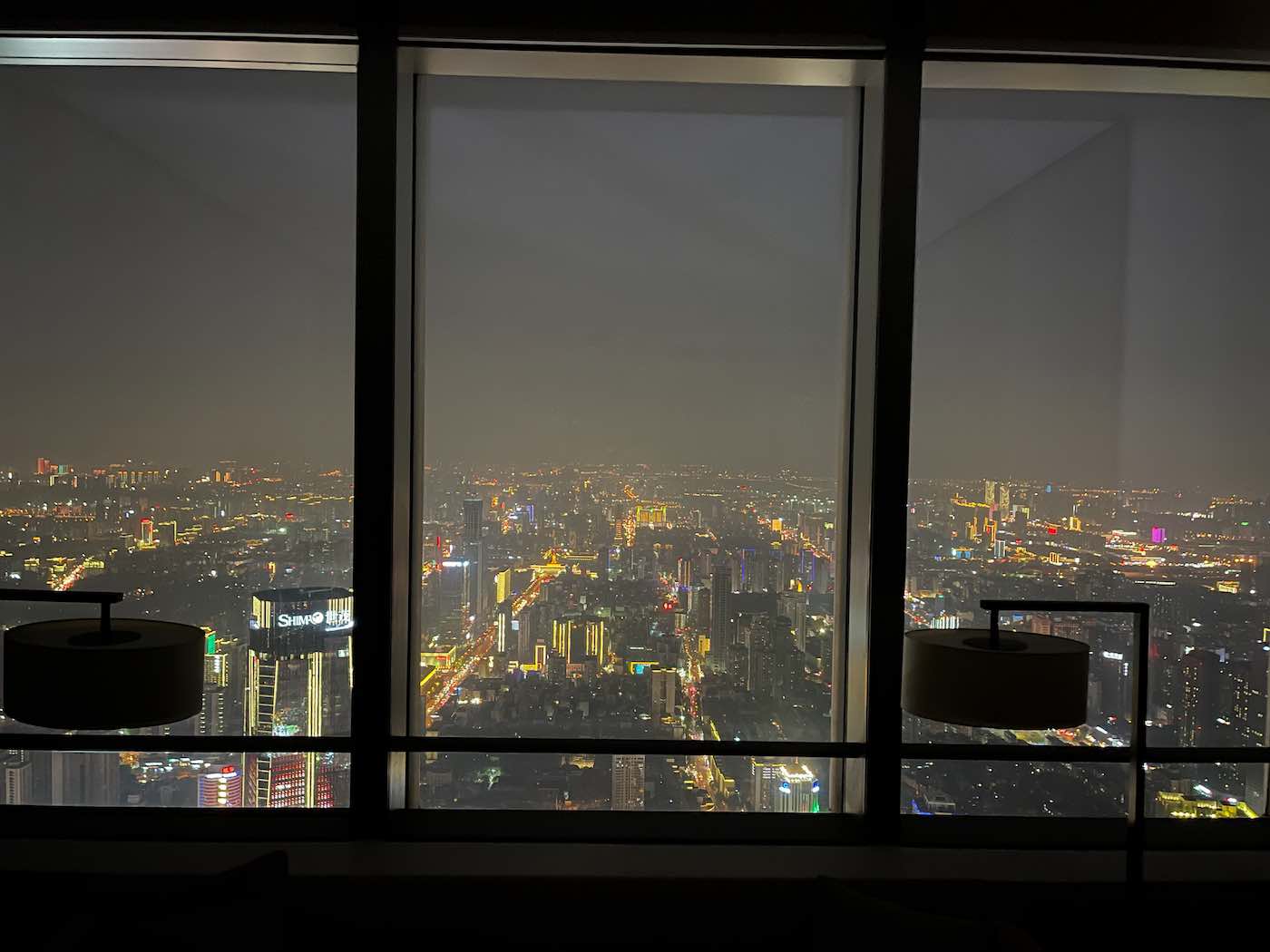 Niccolo Changsha: overnachten in een hotel op 452 meter hoogte 8 Uitzicht op een stadsgezicht 's nachts, gezien door grote ramen, met verlichte gebouwen en straten, en twee silhouetlampen op de voorgrond. -copyright-moving-jack.com
