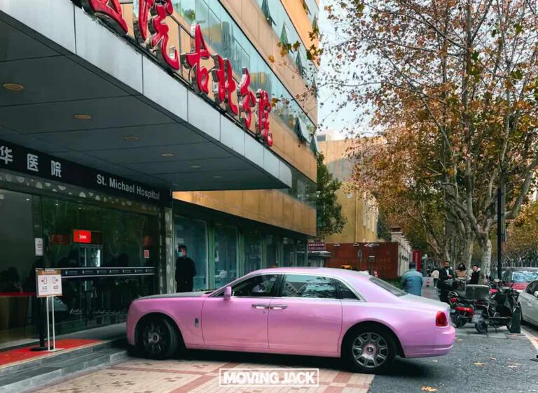 A pink luxury car is parked on a curb outside a building with a sign reading "st. Michael hospital. " there are people nearby, and trees with autumn leaves are visible in the background. This scene could easily be part of an article titled, "what is it like to live in china? The complete guide.