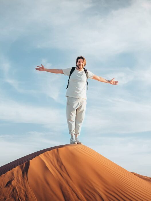 Ultimate guide to the desert in oman: wahiba sands 26 dune bashing in oman Standing at the forefront of a vast sand dune, a person with outstretched arms embraces the elements under a cloudy sky.