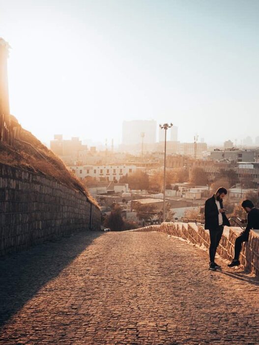 21 unique things to do in erbil 20 Two people sit on a stone wall along a sloping cobblestone path, overlooking erbil's cityscape at sunset, enjoying one of the top things to do in this vibrant destination.