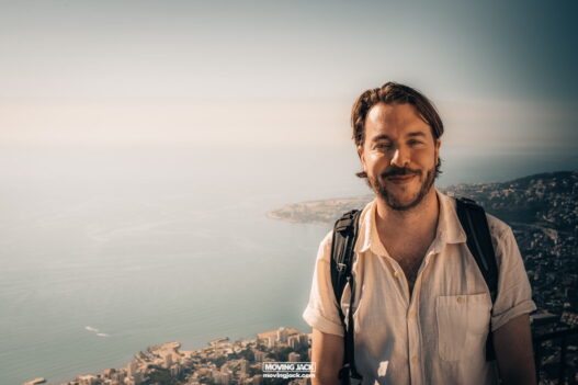 A person with a backpack smiles in front of a scenic coastal view under a clear sky.