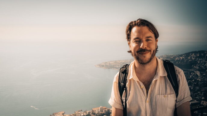 A person with a backpack smiles in front of a scenic coastal view under a clear sky.
