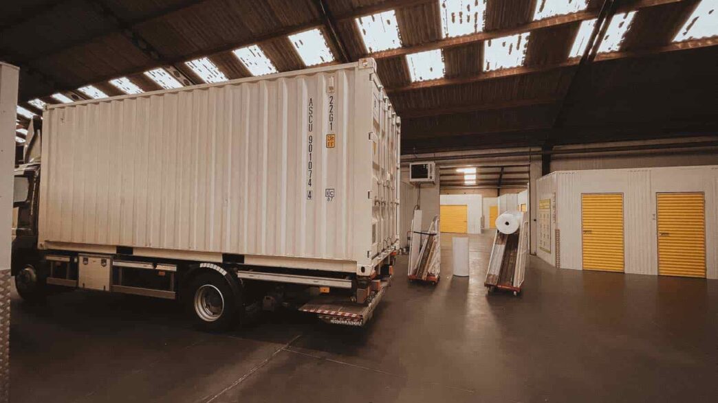 Een witte scheepscontainer op een vrachtwagen staat in een loods met zichtbare dakpanelen en gesloten gele opslageenheden. Het doet me denken aan mijn ervaring toen ik naar een ander land verhuisde.