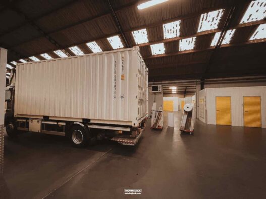A white shipping container on a truck sits inside a warehouse with exposed roof panels and closed yellow storage units, reminding me of my experience of moving to another country.