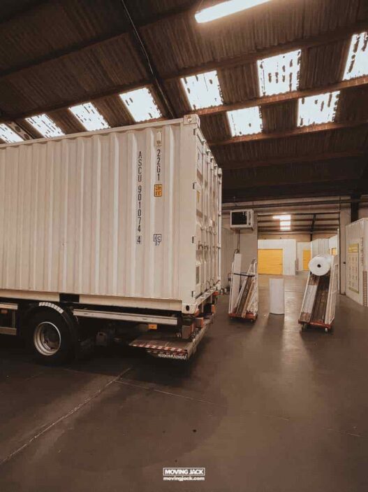 Moving to a new country: the complete checklist 29 travelling with cats A white shipping container on a truck sits inside a warehouse with exposed roof panels and closed yellow storage units, reminding me of my experience of moving to another country.