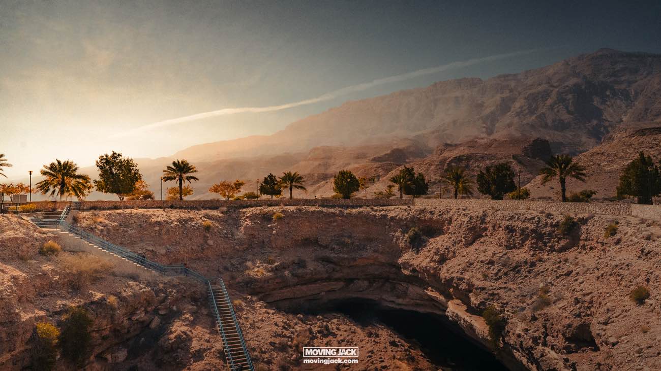 What to see in oman: the 12 sights that will make you love it 20 what to see in oman A scenic view of bimmah sinkhole in oman, featuring a clear blue water hole surrounded by rugged mountains, palm trees, and a wooden pathway under a hazy sky.