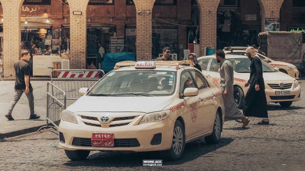 Visiting erbil & iraq for the first time: expert guide + tips 8 first time A white taxi is parked on a cobblestone street in front of a market in erbil. A man wearing traditional clothing is approaching the taxi, while other people and another taxi are seen in the background. This scene captures the essence of daily life for those considering renting a car in erbil.