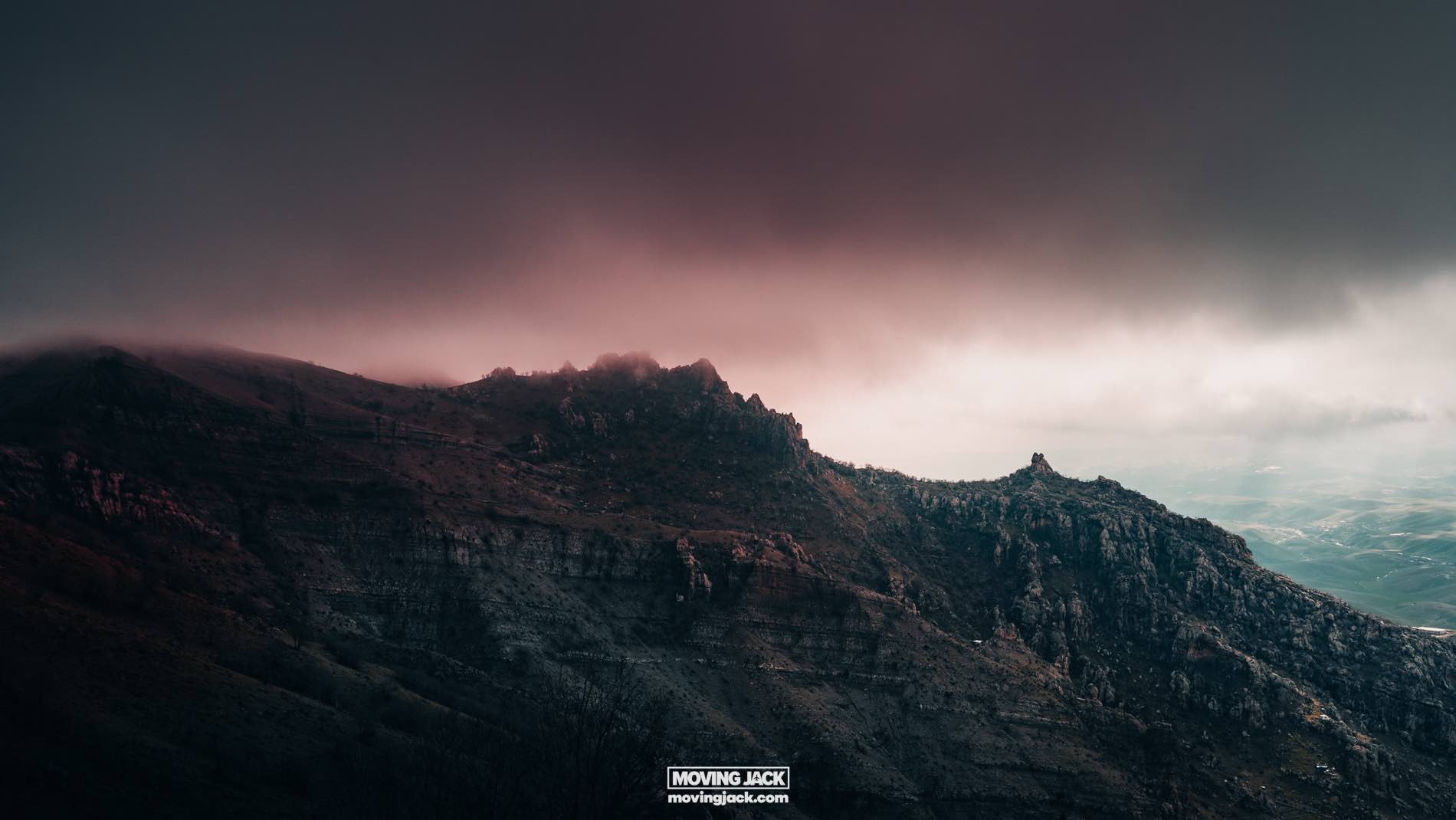A rugged mountain range under a dramatic, cloudy sky with varying shades of pink and gray, reminiscent of the zagros mountains. Text at the bottom reads "moving jack" with a website link.