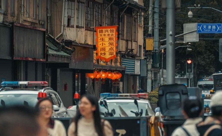 A busy street scene captures the dynamic energy of shanghai, with blurred pedestrians in the foreground and police cars ensuring safety. Lanterns hang gracefully outside a building, while a sign featuring chinese characters adds to the city’s vibrant charm. -copyright-moving-jack. Com