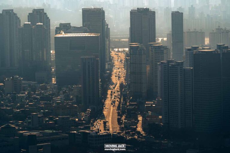 Aerial view of a cityscape with high-rise buildings lining a central road under a hazy sky, offering a glimpse of urban life that makes you wonder what it would be like moving abroad for 3 months to explore the towering architecture and vibrant streets. -copyright-moving-jack. Com