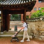 A man, living in seoul, pushes a baby stroller under a traditional korean-style gate, surrounded by stone walls and trees. A blue sign with korean writing stands to the left of the entrance. -copyright-moving-jack. Com