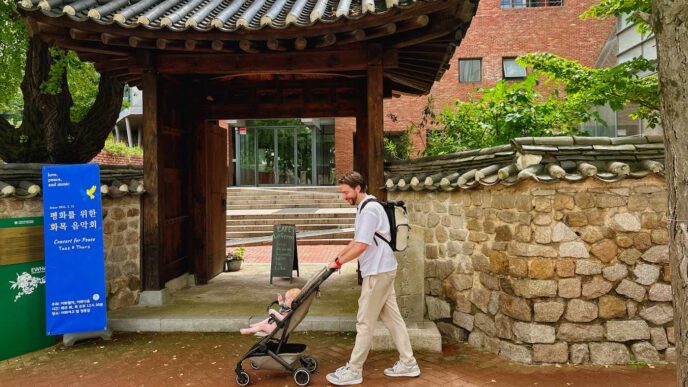A man, living in seoul, pushes a baby stroller under a traditional korean-style gate, surrounded by stone walls and trees. A blue sign with korean writing stands to the left of the entrance. -copyright-moving-jack. Com