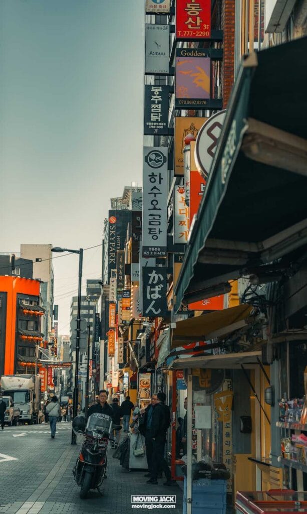Drukke stadsstraat in Seoul met voetgangers en een motorfiets die door kleurrijke verticale winkelborden in verschillende talen slingert. Gebouwen staan aan beide kanten onder een heldere hemel, wat een glimp biedt van waar je kunt verblijven in deze levendige omgeving. -Copyright-moving-jack.com