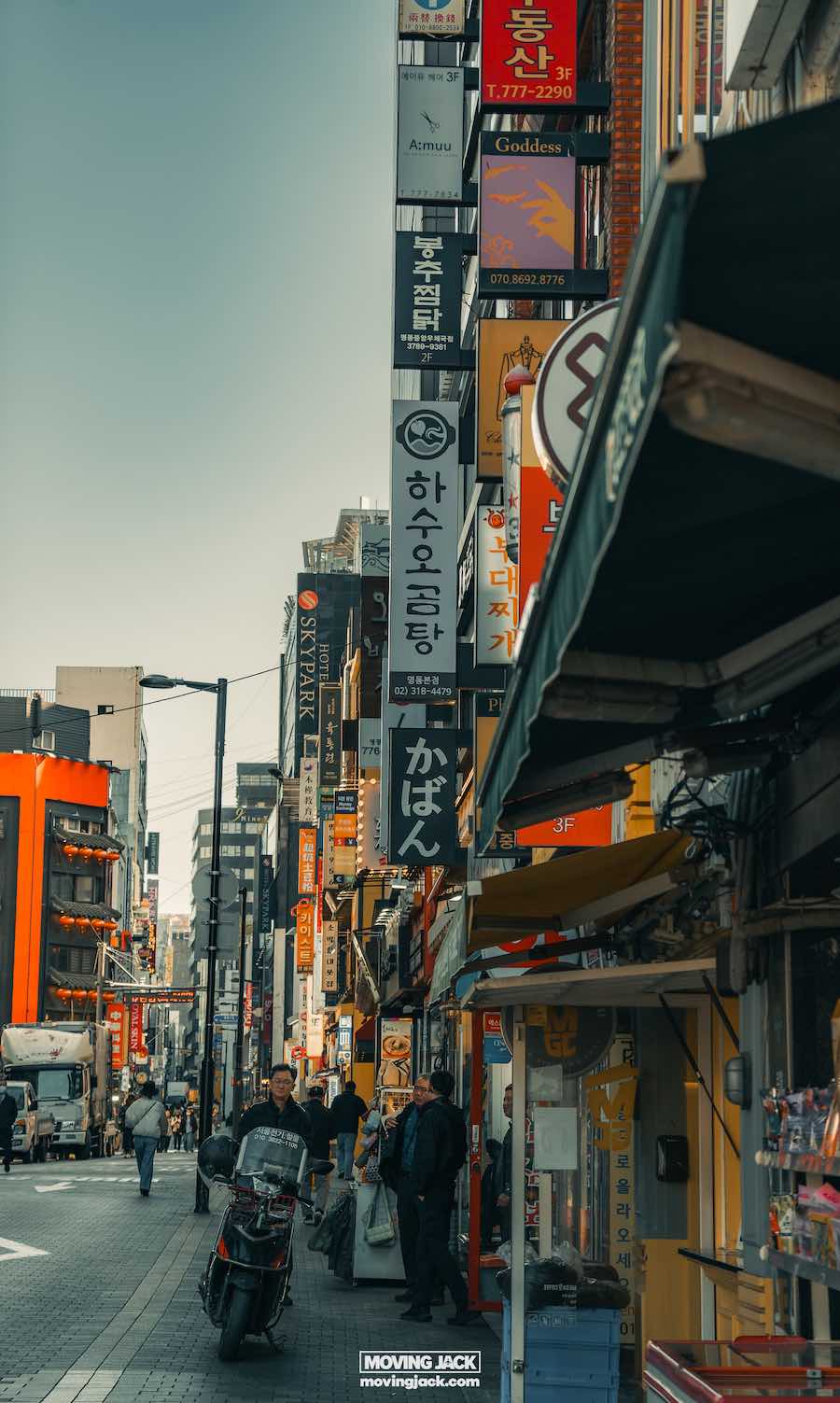 Waar te verblijven in Seoul: beste hotels + reisgids voor gebieden 7 waar te verblijven in Seoul Drukke stadsstraat in Seoul met voetgangers en een motorfiets die door kleurrijke verticale winkelborden in verschillende talen slingert. Gebouwen aan beide kanten onder een heldere hemel, die een glimp bieden van waar te verblijven in deze levendige omgeving. -copyright-moving-jack. Com