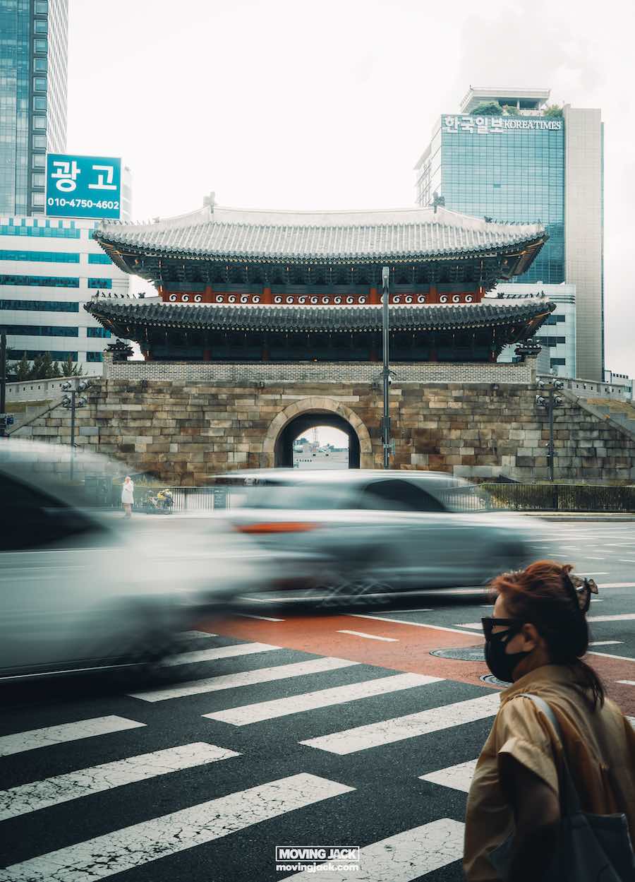 Wat te doen in Seoul: 13 unieke dingen om te doen + wat je beter kunt overslaan! 35 dingen om te doen in Seoul Een persoon met een masker steekt een straat over met wazige auto's die voorbij razen voor een historische Koreaanse poort en moderne gebouwen, en legt de levendige mix van traditie en innovatie vast - ideaal voor degenen die zich afvragen waar ze in Seoul moeten verblijven. -copyright-moving-jack. Com