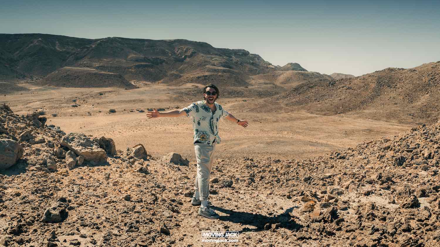 Person standing on rocky desert terrain, wearing a patterned shirt and sunglasses, with arms wide open. Mountainous landscape in the background under a clear sky. -copyright-moving-jack. Com