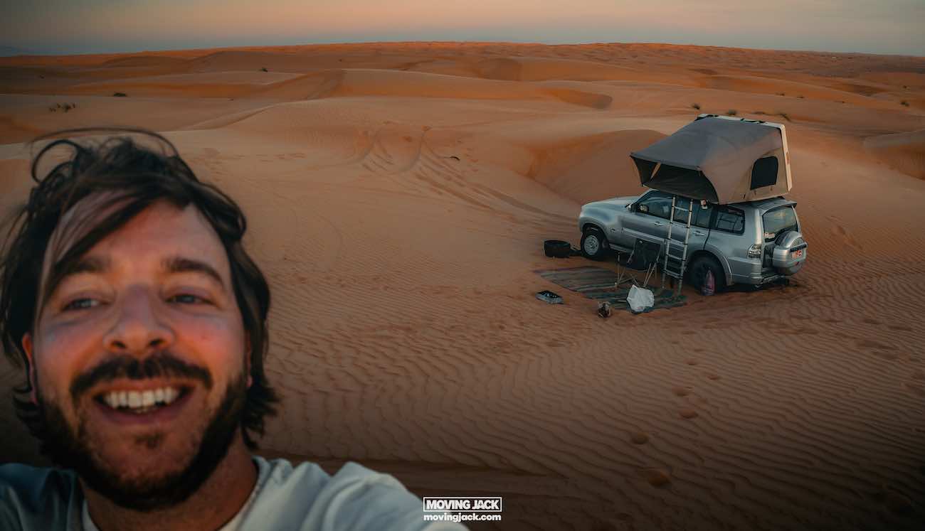 Dune bashing in oman: all you need to know + tours 7 dune bashing in oman A man taking a selfie in the desert after an exhilarating day of dune bashing in oman, stands next to his parked suv featuring a rooftop tent. The sandy landscape stretches into the distance under a dusky sky.