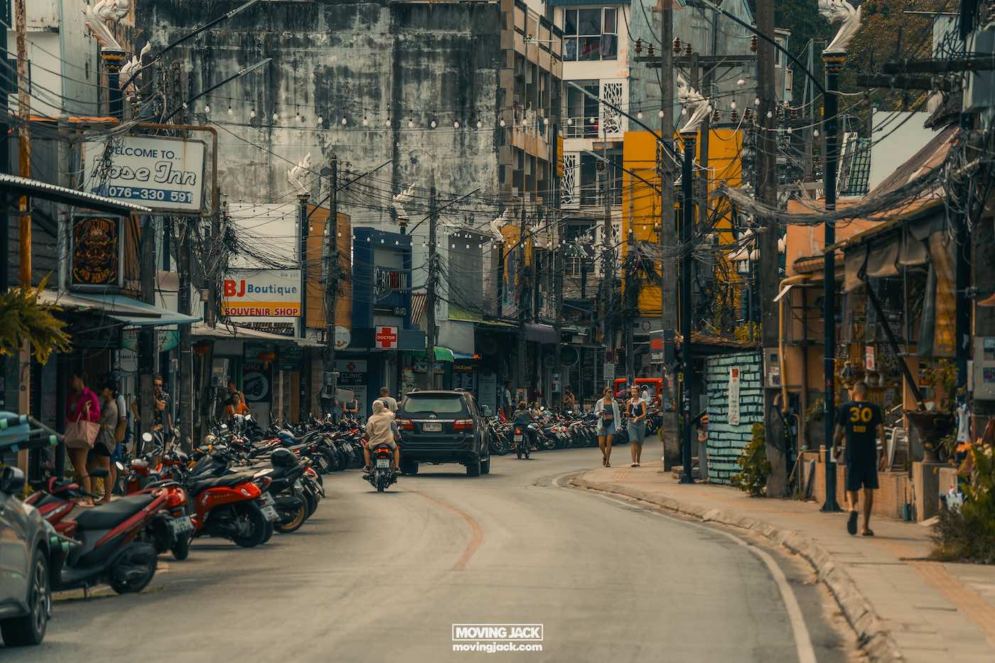 Waarom Phuket Thailand bezoeken? 10 onverslaanbare redenen om te gaan 4 redenen om Phuket te bezoeken Drukke straatscène in Phuket met aan beide kanten geparkeerde motoren, een paar auto's, winkels met levendige bewegwijzering en wandelende mensen. Gebouwen staan dicht op elkaar, met boven je hoofd zichtbare elektriciteitskabels. Het is een momentopname van waarom je Phuket moet bezoeken vanwege de bruisende charme en levendige sfeer. -copyright-moving-jack. Com