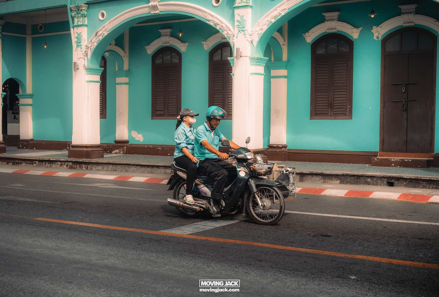 Waarom Phuket Thailand bezoeken? 10 onverslaanbare redenen om te gaan 12 redenen om Phuket te bezoeken Twee mensen op een motor rijden langs een turquoise gebouw met gebogen ramen en deuren, wat de levendige essentie van waarom Phuket bezoeken vastlegt. -copyright-moving-jack. Com