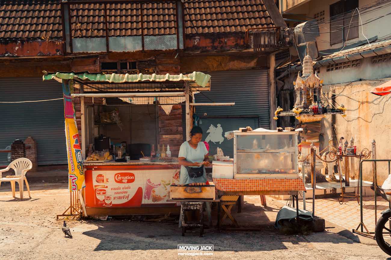 Waarom Phuket Thailand bezoeken? 10 onverslaanbare redenen om te gaan 10 redenen om Phuket te bezoeken Een straatverkoper staat bij een eetkraam onder een provisorisch dak en bereidt eten - een typisch tafereel dat antwoord geeft op de vraag waarom je Phuket moet bezoeken. Er is een gesloten luik en een geestenhuis in de buurt, wat de unieke charme van het eiland vastlegt. Er staat een plastic stoel aan de zijkant. -copyright-moving-jack. Com