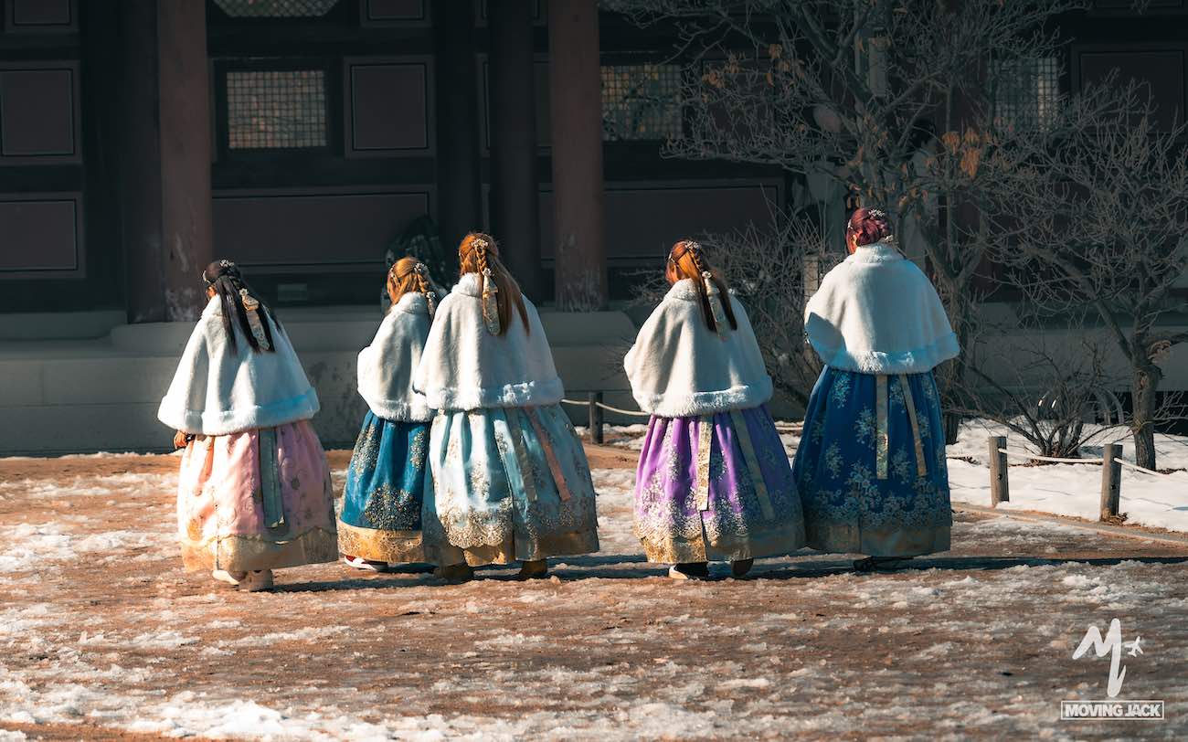 Vijf vrouwen in kleurrijke hanbok-jurken met witte sjaals lopen samen over een besneeuwd pad bij een traditioneel gebouw, waarmee ze de charme vastleggen van het ontdekken van accommodaties in Seoul tijdens de winter.