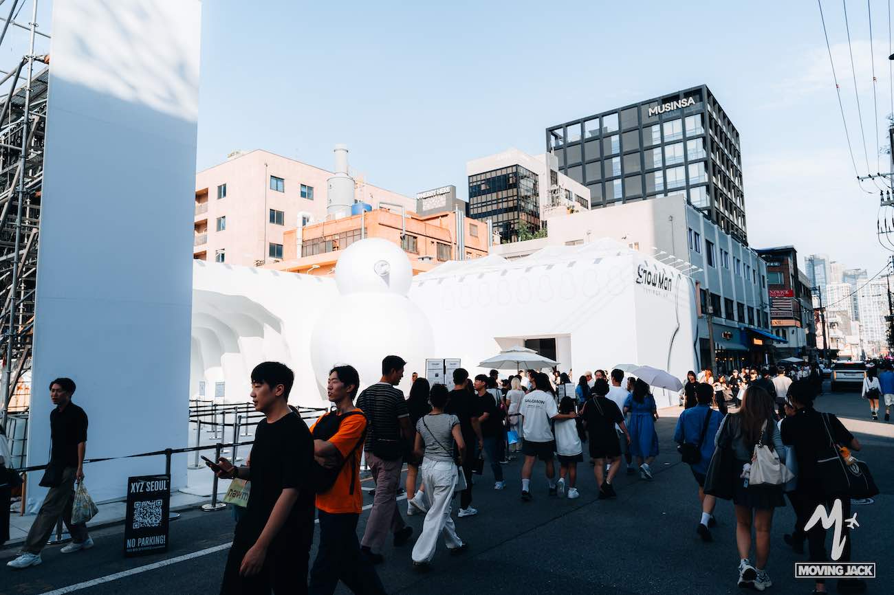 Een menigte loopt langs witte opblaasbare sculpturen en moderne gebouwen in een zonnige stadsstraat en geeft een glimp van de levendige sfeer terwijl je op zoek bent naar een accommodatie in Seoul.