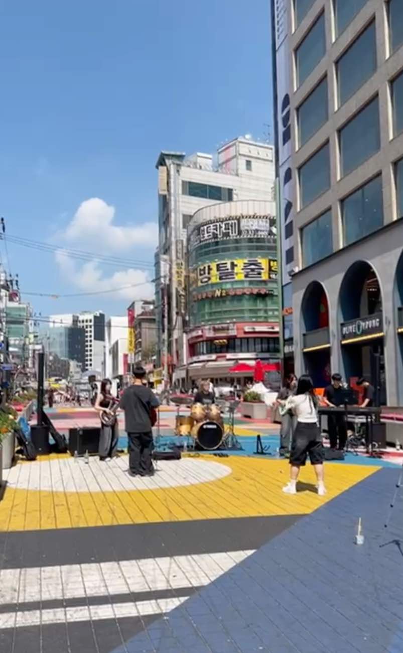 Straatmuzikanten treden op op een kleurrijke stoep in de stad, met hoge gebouwen en winkels op de achtergrond.