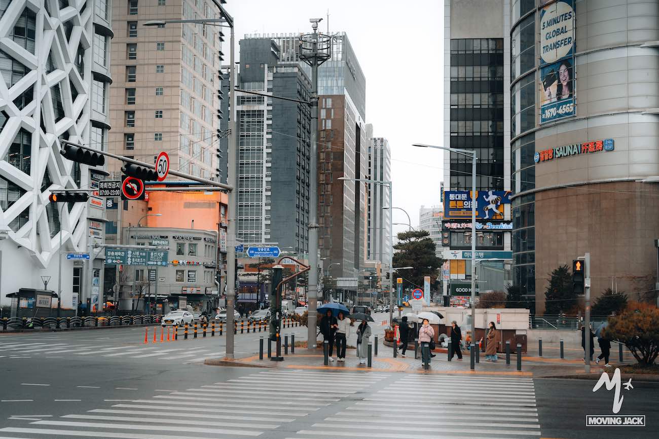 Mensen met paraplu's steken op een regenachtige dag een drukke stadsstraat over op een kruispunt, omringd door hoge gebouwen.