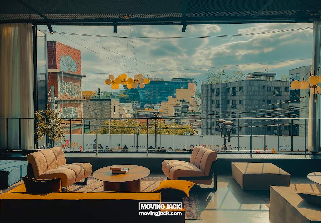 Reisroute Itaewon 2025: de beste eetgelegenheden, drankjes en uitgaansgelegenheden 8 Itaewon Moderne loungehoek met banken en ronde tafels met uitzicht op een stedelijk landschap door grote ramen; heldere verlichting en zichtbare graffiti. -copyright-moving-jack.com