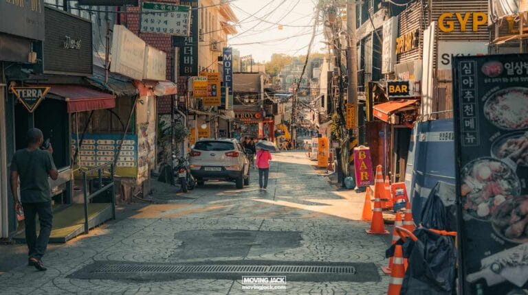 A narrow urban street in itaewon features various shops, signs in korean, orange traffic cones, a few pedestrians, and parked cars. The atmosphere is busy and bustling. -copyright-moving-jack. Com