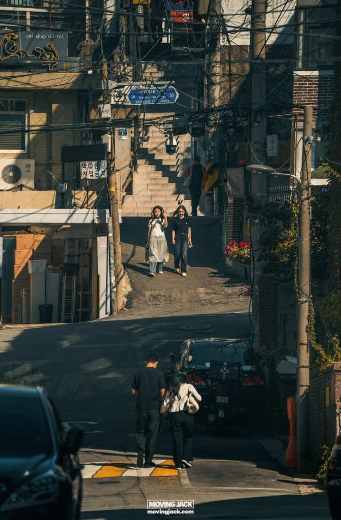 Reisroute Itaewon 2025: de beste eetgelegenheden, drankjes en uitgaansgelegenheden 15 Itaewon Mensen slenteren door een smalle straat in Itaewon, met trappen op de achtergrond, omringd door gebouwen en elektriciteitskabels. -copyright-moving-jack. Com