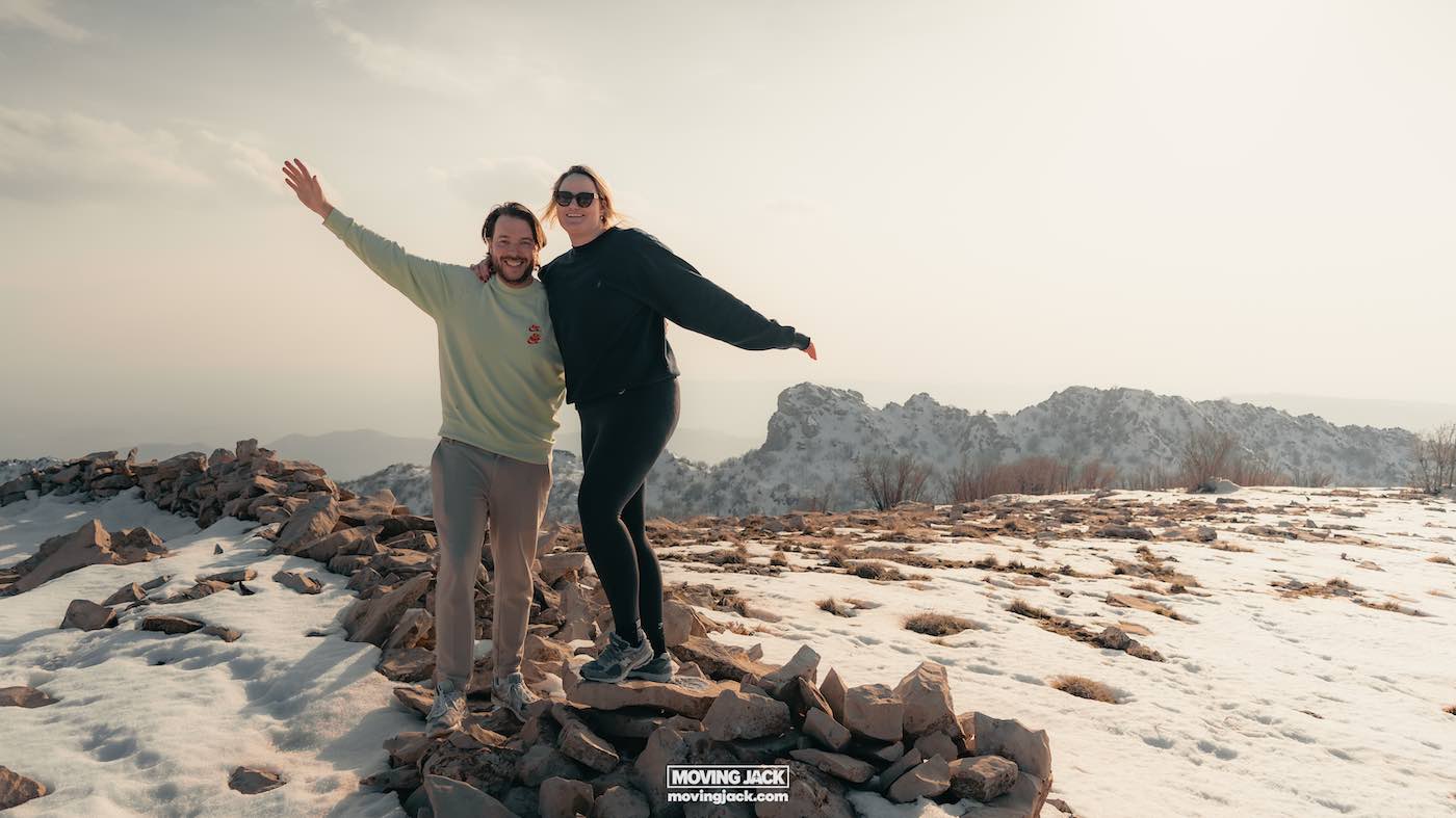 Moving abroad as a couple, two people pose happily on a rocky, snowy hilltop with distant mountains in the background, embracing their shared adventure. -copyright-moving-jack. Com