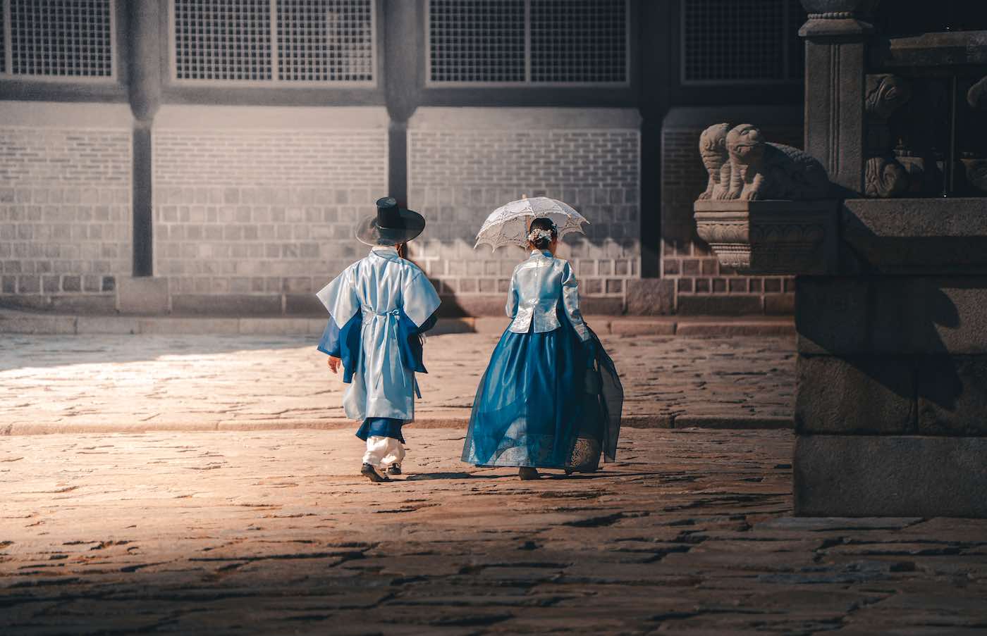 Seoul on a budget: save money in south korea 9 seoul on a budget Two people in traditional korean attire walk along a cobblestone path in south korea. One holds a white parasol. The backdrop features a stone wall and statues. -copyright-moving-jack. Com