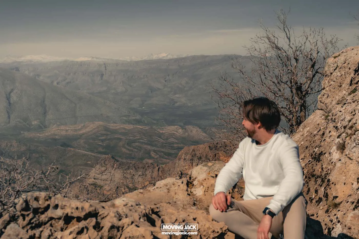 Visiting erbil & iraq for the first time: expert guide + tips 3 first time A person in a white sweater sits on a rocky ledge, experiencing the vast mountainous landscape under clear skies for the first time. Nearby, bare trees stand as silent witnesses to this moment of wonder. -copyright-moving-jack. Com
