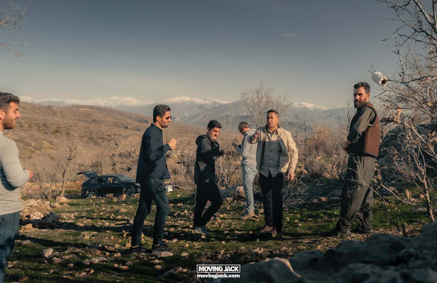 About moving jack 10 about me A group of men, likely first-timers in this rugged terrain, stand outdoors on a rocky landscape with mountains in the background. One man gestures animatedly as the others observe, intrigued. A car is parked nearby, ready for their next adventure. -copyright-moving-jack. Com
