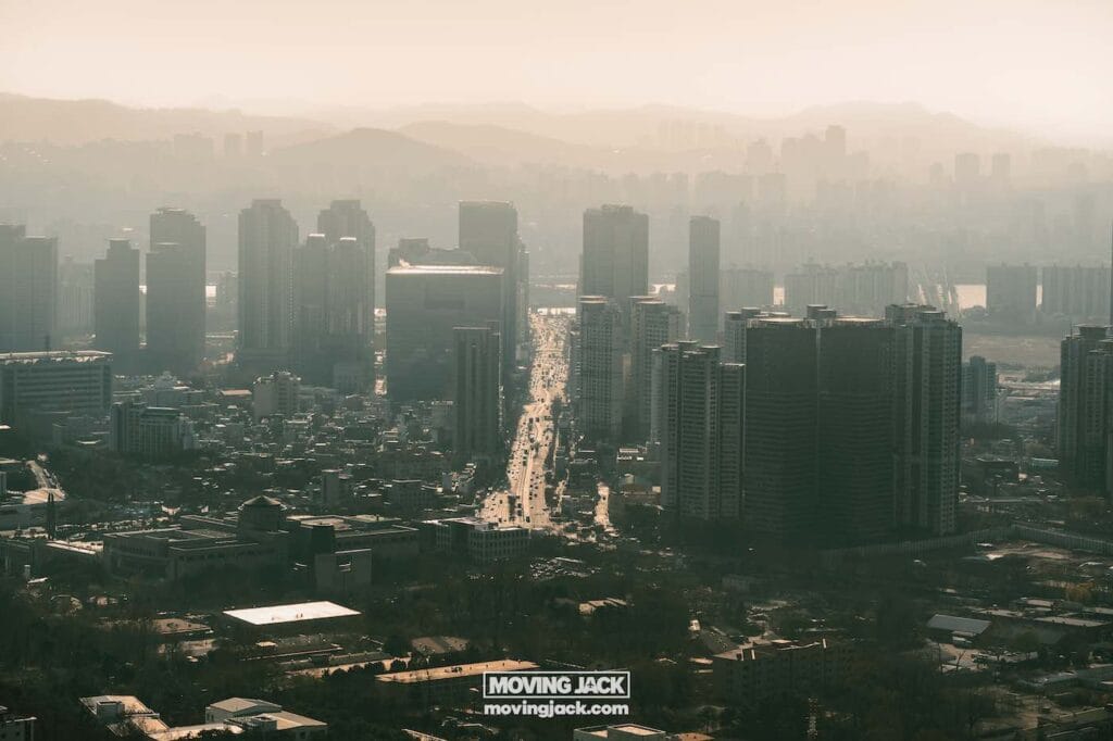 Stadsgezicht met hoge gebouwen langs een hoofdweg. In de verte zijn heuvels zichtbaar onder een wazige hemel, waardoor een onscherpe stedelijke horizon ontstaat. -Copyright-moving-jack.com