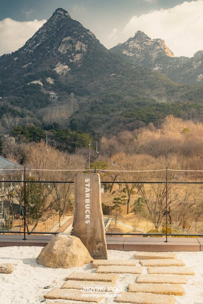 Wat te doen in Seoul: 13 unieke dingen om te doen + wat je beter kunt overslaan! 11 dingen om te doen in Seoul Een houten bord met "starbucks" staat in een natuurlijke omgeving met bergen en bomen op de achtergrond onder een bewolkte lucht. -copyright-moving-jack. Com