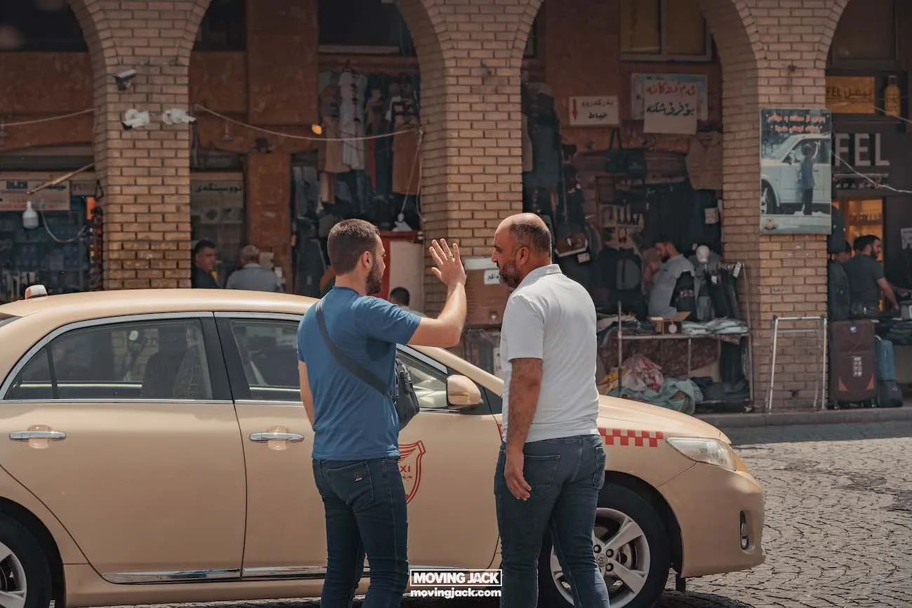 Visiting erbil & iraq for the first time: expert guide + tips 5 first time Two men, appearing as first timers in the bustling city, stand in conversation next to a parked yellow taxi on a cobblestone street, with a lively market area visible in the background. -copyright-moving-jack. Com